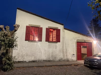 This image depicts a street scene at dusk or night in the city of Caminha, Portugal. The central focus is a single-story, white-washed building with a sloped, terracotta-tiled roof. The building features two windows with open, bright red wooden shutters; both interiors are illuminated with a warm, reddish light. To the right, a large, partially open bright red double door reveals an illuminated interior. A house number, "40," is affixed above the doorframe. The exterior wall has minor discoloration, particularly near the base. A small, grey utility box is mounted on the wall below the right window.
To the left of the building, a mature, gnarled tree with exposed roots and some leafy growth, possibly ivy, extends into the frame. A small, illegible street sign is attached to this tree. The foreground consists of a cobblestone street, which curves slightly, and a narrow, light-colored sidewalk. Scattered dry leaves are visible on both the street and sidewalk.
A dark grey sedan, identifiable as a Renault, is parked on the cobblestone street in front of the building, facing left. Its license plate, "50 28 ZI," is visible. A bright artificial light source, likely a wall-mounted fixture, is positioned above the building's main door, casting a prominent lens flare across the upper right portion of the image. The sky is a deep twilight blue. In the far right background, a tall, dark coniferous tree is silhouetted against the sky. No individuals are present in the scene.
