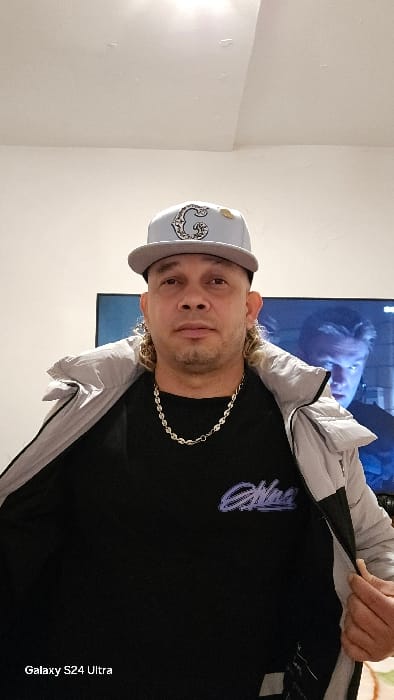 This image captures a male individual indoors within a building located in Real Sitio de San Ildefonso, Spain. The subject is a male of apparent middle age, possessing light skin, facial stubble, and tightly curled, light-colored hair styled as a mullet, visible extending from beneath his headwear. He is wearing a light gray baseball cap with a dark gray brim, featuring an ornate letter "G" embroidery and a small gold-colored pin. Around his neck is a silver-colored chain necklace with oval links. He wears a black t-shirt adorned with a purple-blue graphic design on the left chest area, and an open light gray and dark gray/black puffer jacket. His hands are positioned to hold the front edges of the open jacket. He gazes directly at the viewer with a neutral facial expression. The background features a plain, light-colored wall and ceiling. Behind the individual, to his right (viewer's left), a television screen is visible, displaying a scene that includes a male figure with a distinct blueish glow illuminating his face.