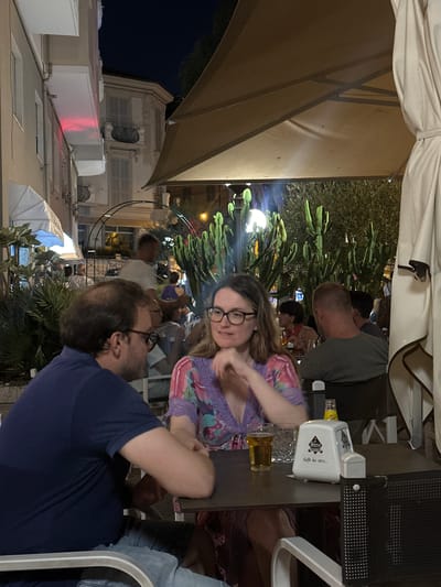 The image depicts an outdoor seating area of a cafe in Ospedaletti, Italy, at night. Two people, a man and a woman, are seated at a table in the foreground, appearing to be in conversation. The man, on the left, has dark hair and wears a dark blue polo shirt. He is looking to his left. The woman, on the right, has lighter hair and wears glasses and a purple and pink patterned top. She is looking down and to her left.
On the table are a glass of beer, an empty glass, a napkin dispenser with "Giena Caffè dal 1869..." printed on it, and a bottle of Schweppes. Behind the couple, other patrons are visible in the background, along with large potted cacti and foliage. Buildings with architectural details, typical of Italian coastal towns, are seen in the distance, illuminated by artificial light. A large beige umbrella provides shade over the seating area. The overall atmosphere suggests a relaxed evening at a cafe.