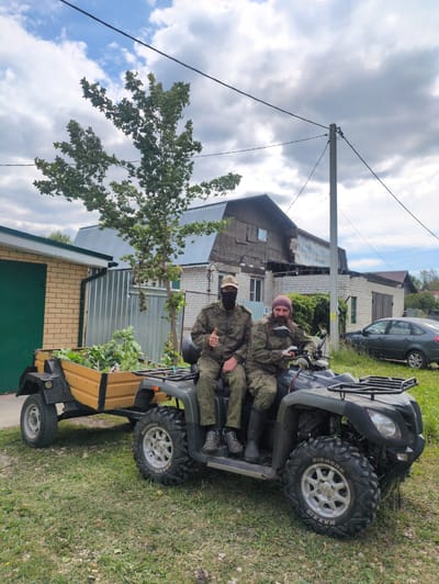 The image shows two individuals in camouflage clothing seated on an all-terrain vehicle (ATV) with a trailer attached. The ATV is parked on a grassy area next to a building with a tin roof. The individuals appear to be in a rural setting, possibly in Великово, Russia, given the context. One person is driving the ATV and is giving a thumbs-up gesture, while the other is seated behind. They are both wearing camouflage uniforms and one of them has a pink beanie and a beard. The weather appears to be overcast, with some clouds visible in the sky. The time of day is likely daytime, given the lighting conditions. There are several buildings visible in the background, including a brick building and what appears to be a house. A grey car is also parked nearby. The ATV has "MAXXIS BIGHORN" written on its tires.