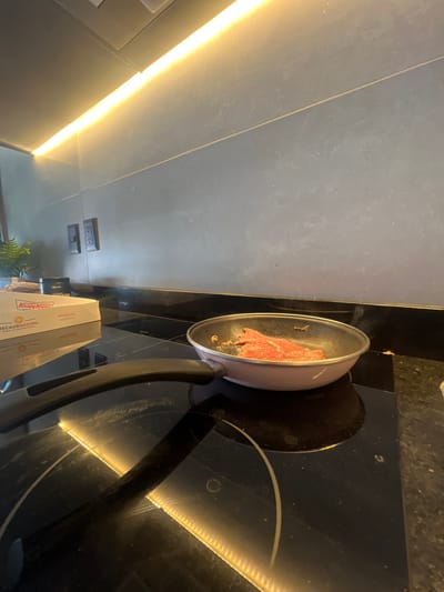 The scene depicts a contemporary kitchen environment located in León, Mexico. In the mid-ground, a light pink frying pan containing several slices of raw or lightly seared red meat, seasoned with visible spices, rests on a black induction cooktop. To the left of the pan, a white Krispy Kreme box, partially showing the word "RECAUDACIÓN," is present on the dark, reflective countertop. The background consists of a dark gray tiled or paneled backsplash wall with two dark electrical outlets mounted. Above the wall, a linear warm-toned LED light strip illuminates the area. A hint of green foliage from a plant is visible on the far left. No individuals are present in the image.