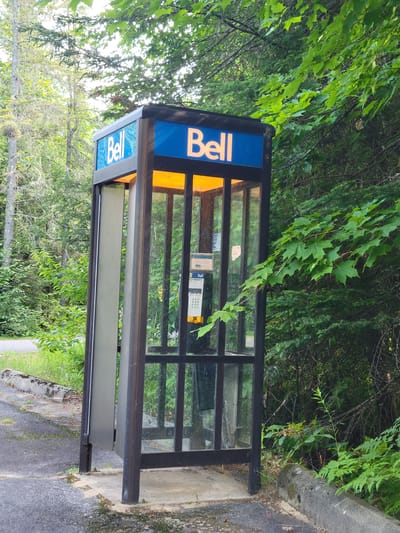 This is a full, eye-level, outdoor shot of a Bell payphone booth situated at a roadside in a wooded area, likely in Shawinigan, Canada, given the prominent "Bell" signage, a Canadian telecommunications company. The payphone booth has a dark metal frame and glass panels, with a blue sign at the top featuring the white "Bell" logo. The interior of the booth is illuminated by a warm orange light from above, and a payphone is visible, with a keypad and screen. A closed door panel is on the left side of the booth, and the right side is open. The ground around the booth is paved with asphalt, with a concrete curb and a heavily vegetated natural environment bordering it. The surrounding area is densely populated with trees and foliage, suggesting a rural or park-like setting. There are no people visible in the image.