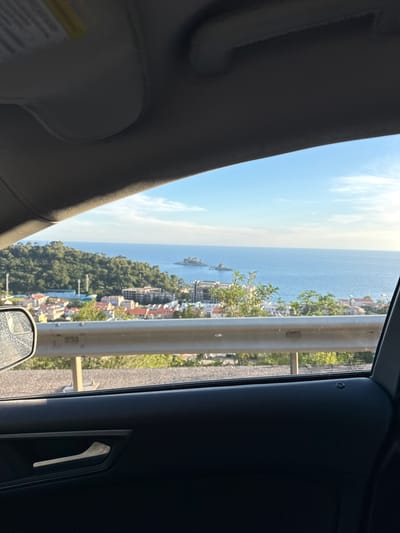 The image is taken from inside a car, looking out the window at a coastal town. The interior of the car is dark, with the door panel and a chrome door handle visible in the foreground. A side mirror with a reflective surface showing dirt and water spots is positioned on the left side of the frame.
Outside the car, a guardrail runs horizontally across the middle of the image, separating the road from the view beyond. Beyond the guardrail, a town with red-tiled roofs and modern buildings is nestled on a hillside. Further in the distance, the blue expanse of the Adriatic Sea stretches to the horizon, dotted with small islands. The sky above is clear blue with a few wispy clouds. Lush green trees and foliage cover the hillsides surrounding the town and reaching down towards the coast. The overall impression is a scenic view of a coastal settlement in what is likely a sunny, temperate climate.
This scene is consistent with the town of Novoselje, Montenegro, known for its coastal location, hilly terrain, and proximity to the sea.