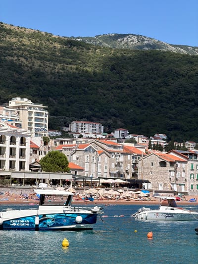 The image shows a view of the Petrovac centar, Montenegro, on a sunny day. In the foreground, two boats are moored in the clear blue water. The boat on the left is white with blue graphics depicting a marlin and the text "RESTORAN ORADA". The boat on the right is also white and has the text "BARACUDA" on its side. Buoys, one yellow and one orange, float in the water.
Behind the boats, a sandy beach is visible, lined with numerous lounge chairs and umbrellas. Several people are scattered across the beach, some sunbathing, others wading in the water. Buildings with red-tiled roofs and stone facades line the waterfront, with modern apartment buildings and hotels rising behind them. The town is nestled against a steep, forested hillside that slopes up to a clear blue sky. The foliage on the hill is dense and green, with patches of lighter green indicating sparser vegetation or rockier terrain higher up. The overall impression is one of a vibrant coastal resort town.
