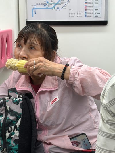 An Asian woman is sitting on a subway or train, holding a cob of corn in her right hand and taking a bite. She is wearing a pink jacket over a red shirt, and has a beaded bracelet on her left wrist. A cell phone with a decorative case is tucked into her jacket pocket. To her left, a backpack with a camouflage pattern is visible. In the background, a map of the city's subway system is displayed on the wall, indicating the location is within the city of Longxi Sub-district, China.