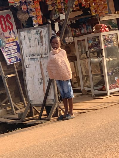A child, wearing a pink sweater and denim shorts, lies asleep on what appears to be a wooden structure outside a roadside shop. The shop displays various goods, including snacks and drinks. Adjacent to the wooden structure is a sign with text in English including, "PLACE HERE," "WITHDRAW," "DEPOSIT," "PAY BILL," and "AIRTIME." The image is tilted, showcasing the street and the exterior of the commercial space.