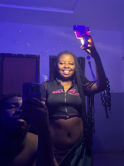 The image captures two individuals in an indoor setting in Gbongan, Nigeria, under dominant blue-purple ambient lighting. A female, positioned centrally, with dark skin, long braided hair, and a nose piercing, is smiling broadly with her tongue extended. She wears a black cropped top featuring pink lettering and a front zipper, exposing her abdomen, and dark bottoms. Her right arm is raised, holding a translucent, toy-like blaster that emits a bright orange-yellow light from its front, appearing purple under the ambient light, with a small purple charm dangling from it. In the lower-left foreground, partially visible, is a male with dark skin and short dark hair, holding a dark-cased smartphone with a triple camera array, indicating he is taking the photograph, likely a mirror selfie. The background displays light-colored walls speckled with numerous reflective points and two dark, rectangular openings.