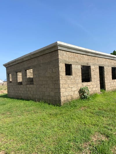 The image shows a partially constructed building made of cinder blocks, with open window and door frames. The building has a decorative concrete trim along the top edge. It is situated in a grassy, undeveloped area with a clear blue sky overhead. In the background, another building with a blue roof is visible. The scene appears to be in Kuson, Nigeria, an area characterized by its natural landscape and ongoing construction projects.