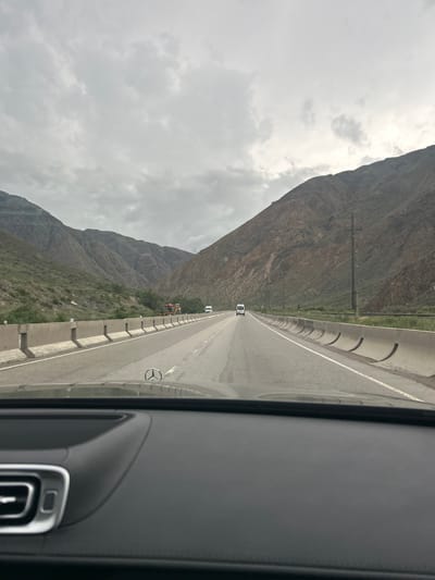 The image shows a view from the interior of a car, looking out at a highway through a mountainous landscape. The road ahead is paved and marked with white lines. On the left side of the road, a concrete barrier separates the highway from a steep, vegetated mountainside. On the right side, the highway is bordered by a similar concrete barrier and then by another steep, rocky mountainside. Several vehicles are visible on the highway: a white van is in the distance, and a yellow construction vehicle is on the shoulder of the road. The sky is overcast with gray clouds. The dashboard of the car is visible in the foreground, with a Mercedes-Benz logo on the steering wheel. The overall scene suggests a journey through a remote, mountainous region. The location is identified as Unknown, Kyrgyzstan.
