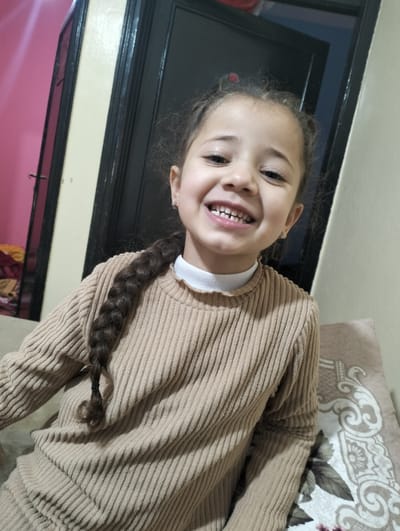 A smiling young girl is depicted indoors. She is wearing a brown, ribbed sweater with a white collar. Her hair is braided and she has earrings. The background features a door with a dark frame and beige and pink walls. A portion of a patterned cushion is visible in the lower right.
Ouaouizert, Morocco is not apparent in the image.