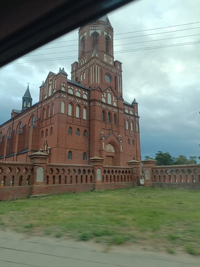 The image captures a large, red brick church in Kuczyn, Poland, under a cloudy sky. The church features a tall, ornate tower with a clock face. A low brick wall with decorative openings surrounds a grassy area in front of the church. Power lines are visible in the sky above the church. The image appears to have been taken from a moving vehicle, as indicated by the motion blur in the foreground and the oblique angle.