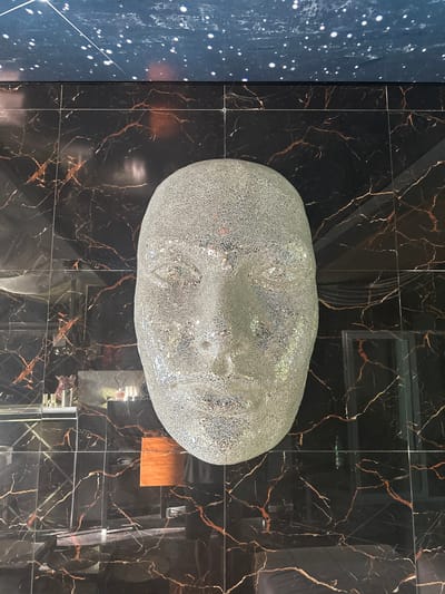 A large, glittery, human-like head sculpture is the central focus of this image, mounted against a wall covered in black marble-patterned tiles with thin, coppery veins. Above the tiled wall is a dark blue ceiling adorned with white speckles resembling stars. The sculpture's surface reflects light, giving it a shimmering appearance. Below the head sculpture, the tiled wall continues, revealing reflections of objects within the room, including what appear to be shelves with bottles and other decorative items, as well as a glimpse of a person's legs and torso wearing dark clothing. The overall environment suggests an indoor, possibly artistic or decorative setting. The image was taken at the hotel Splendid Conference & Spa Resort in Bečići, Budva, Montenegro.