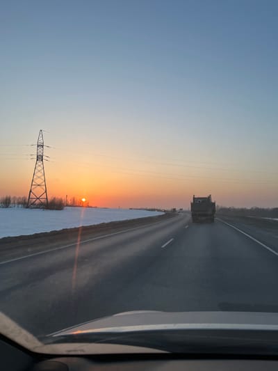 The image depicts a winter highway scene at either sunrise or sunset, viewed from inside a moving vehicle, located in or near Shelepino, Russia. The sky transitions from light blue at the zenith to vibrant orange and yellow hues at the horizon. A prominent, bright orange sun is low on the horizon, positioned left of center.
The foreground shows the lower section of a car's windshield and dashboard, with a reflection of the sun visible. The asphalt highway, appearing dark and possibly wet, recedes into the distance with white solid and broken lane markings. On the left side of the highway, a wide expanse of snow-covered ground is visible, dotted with sparse, leafless trees and bushes. A tall, metallic lattice-type power line tower stands prominently on this snowy plain, with power lines extending horizontally across the mid-ground.
In the right lane of the highway, a dark-colored dump truck with an open or slightly raised bed is observed moving away from the camera. Additional vehicles are visible as distant silhouettes further down the road. No individuals are explicitly depicted.