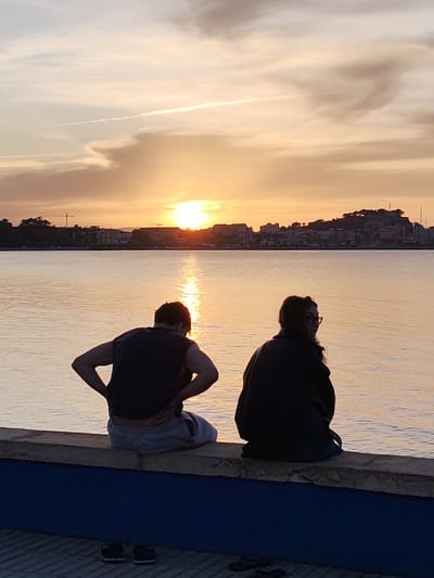 Two individuals are seated on a concrete ledge with a painted blue base, overlooking a body of water reflecting a sunset. To the left is a person with short dark hair, wearing a dark sleeveless shirt and light-colored shorts. Their hands are on their hips. To the right is a person with long dark hair and glasses, wearing a dark jacket. Both are facing the water. The sunset is visible in the distance, over a cityscape with buildings and a small hill. A light contrail stretches across the sky. Pairs of shoes are visible on the ground in front of the ledge. The location is Dénia, Spain, as indicated by the cityscape.