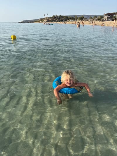 A child with blond hair is floating in the water, wearing a blue flotation device. The water is clear enough to see the sandy bottom. In the background is a beach in Nahariah, Israel, with buildings and people. A yellow buoy is visible in the water near the child.