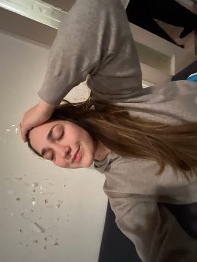 A woman with closed eyes leans her head on her hand. She wears a gray sweatshirt and her light brown hair is parted on the side. Small, shiny, semi-transparent objects are scattered across the white surface in front of her. A door frame and black-clad legs are visible in the background.
