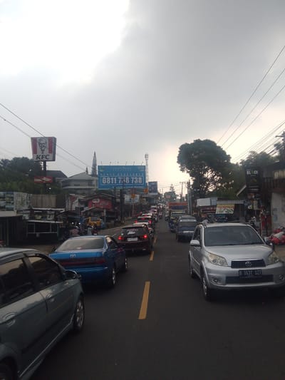 A medium shot captures a congested street in Cisarua, Indonesia, under an overcast sky. The scene is dominated by a traffic jam of various vehicles, including cars, trucks, and motorcycles, all moving slowly on a two-lane road. The road is marked with yellow lines.
On the left side of the road, a large KFC sign with Colonel Sanders' image is prominently displayed. Below it are several shops and stalls, some appearing to be for food vendors. Further down the street, a blue Toyota sedan is visible, followed by other vehicles.
On the right side of the road, a silver Toyota SUV is in the foreground, facing the camera. Further down, more cars and a large billboard advertising "SPACE AVAILABLE" with a phone number are visible. Another billboard for "BLACK" coffee and a price list is also present. The buildings along the street are of varying sizes and styles, some appearing to be commercial establishments. Trees and vegetation are visible in the background, suggesting a somewhat suburban or semi-rural setting within the city. The overall atmosphere suggests a typical busy day with heavy traffic.