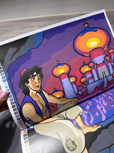 The media file shows a close-up, high-angle shot of a coloring book spread featuring the character Aladdin. Aladdin is depicted in a stylized, colorful illustration. He is wearing his signature purple vest and red vest. The background is a vibrant purple and red gradient, with a fantastical cityscape or palace in shades of purple and blue. The illustration appears to be in progress, with the coloring done in a flat, bold style.
Aladdin's expression is one of contemplation or perhaps awe, as he looks upwards towards the fantastical architecture. His body is positioned as if he is sitting or leaning back.
At the bottom edge of the coloring book page, there are visible lines of letters and numbers, indicating this is a numbered or lettered coloring book. Partially visible letters include 'c', 'd', 'e', 'h', 'j', 'k', 'm', 'n', 'p', 'q', 'r', 't', 'w', 'x', 'y', 'z', 's', and numbers '5', '6', '7', '8', '9', '0'. A portion of a finger with light purple nail polish is visible in the lower right corner, holding down the edge of the page. The overall impression is a bright, fantastical scene rendered in vivid colors. The location context provided is Łódź, Poland.