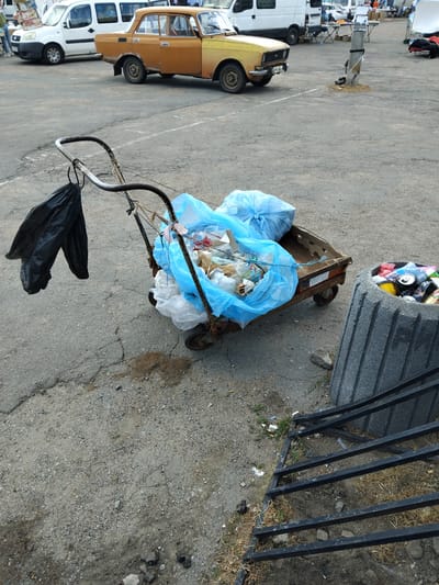 The image depicts an outdoor scene in Chudniv, Ukraine, likely a market or an open area. In the foreground, a rusted handcart is filled with garbage bags, primarily blue and white, along with loose trash. Black trash bags are also hanging from the handle of the cart. To the right, a large, grey, concrete trash receptacle is overflowing with discarded cans and other refuse. The ground is paved with asphalt, showing signs of wear and cracks, with scattered debris and dry grass. In the background, several vehicles are parked, including a white van and a yellow sedan. The environment appears to be a public space, possibly a street or a parking lot, with a general sense of disarray and litter.