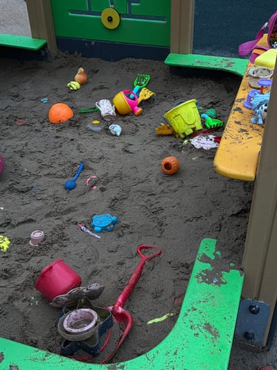 The image depicts a close-up view of a sandbox filled with dark, granular sand. The sandbox contains a variety of scattered children's toys, including buckets, shovels, a watering can, various plastic molds shaped like animals and objects, and a red plastic rake. Some of the toys are partially buried in the sand. The sandbox appears to be part of an outdoor playground structure, with glimpses of green and yellow plastic components of the structure visible at the edges of the frame. The overall scene suggests a recently used play area. There is no indication of the location in the city of Фролы, Russia, within the image.