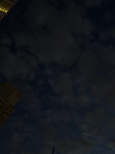 An overhead view looking up at a dark, cloudy sky with two buildings visible. The building on the left is light brown with multiple floors and rectangular windows. The building on the far left is mostly obscured but appears to be a taller structure with illuminated windows. Power lines are visible in the lower right quadrant of the image. The scene is set in Shushary, Russia.