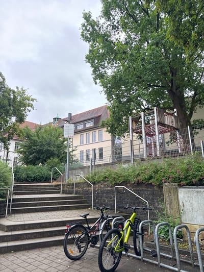 In Bietigheim-Bissingen, Germany, a cloudy day frames a scene featuring a building with a red-tiled roof, multiple windows, and a distinctive green dome. In front of the building is a fenced-off playground area with a wooden and metal structure. A set of stone stairs leads up to a paved area where two bicycles are parked at a bike rack. The foreground shows the bicycles and the bike rack, with the stairs and building in the background. The environment is urban, with trees and greenery adding to the scenery.