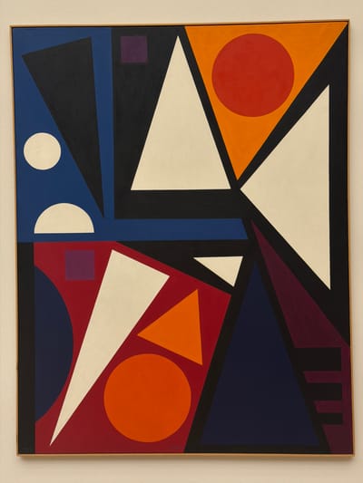This is a forensic, matter-of-fact description of an abstract geometric painting. The painting is framed in light wood and mounted on a neutral-colored wall, suggesting it is on display in a gallery or museum. The artwork features a vibrant composition of interlocking geometric shapes in bold colors, including red, orange, blue, black, white, and purple. There are triangles, circles, squares, and semi-circles arranged in a dynamic and balanced manner. The image does not depict any people, actions, or interactions. The setting is a sterile exhibition space, typical of a museum in Basel, Switzerland, such as the Kunstmuseum Basel, which houses a significant collection of modern and contemporary art.