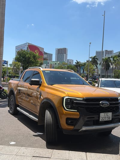 An orange Ford Ranger pickup truck is parked on a city street in Quy Nhon Ward, Vietnam. The truck has a black grille with a Ford logo and a license plate that reads "77C 227.58". There are also two white surgical masks on the ground in front of the truck. In the background, there are several buildings, palm trees, and a large billboard with a red hammer and sickle symbol. The sky is blue with a few white clouds. The scene appears to be a sunny day.