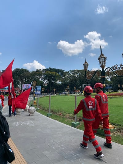 Two men wearing red uniforms with the logo "PMI" are walking on a paved sidewalk in front of a grassy area. The sidewalk has white chalk markings and a street lamp with a golden top. In the background there are trees, a red flag on a pole, and a blue sky with white clouds. The scene is in Malang, Indonesia, at the coordinates -7.977 latitude and 112.634 longitude.