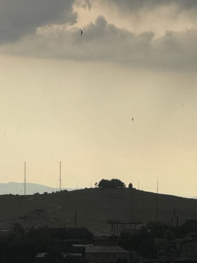 The image, taken in Abovyan, Armenia, shows a landscape with a cloudy sky. Two planes are visible in the sky; one is closer to the bottom left, flying near a large cloud formation, and another is further away in the center of the frame. The sun appears to be setting, casting a warm, hazy light across the scene, suggesting it's either early morning or late afternoon.

The foreground on the right side of the image is dominated by a dark, silhouetted hillside. On the hillside, structures resembling houses or buildings are dimly visible, as are several tall, slender poles, possibly utility poles or communication towers. Trees are also scattered across the hillside. The sky itself is a mix of light gray and pale yellow, with some darker, more defined clouds on the left. The overall atmosphere is somewhat muted and serene. No text is visible in the image.