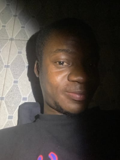 A close-up shot of a dark-skinned man laying down. The man is on a tiled floor with a repeating pattern. He is wearing a black shirt with a blurred design on the right shoulder. His head is slightly tilted, facing the camera with his eyes partially open. The lighting is low, casting shadows on his face and hair, which is short and closely cropped.