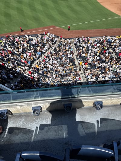 The image depicts an aerial view of a baseball game in progress at Petco Park in San Diego, California, United States. A crowd fills the stadium seating adjacent to the field, comprised of adults and children, many wearing baseball jerseys. Near the field a person in red garb is seen. The foreground shows stadium seating, a transparent partition, and cup holders. There is clear sunlight, casting shadows.