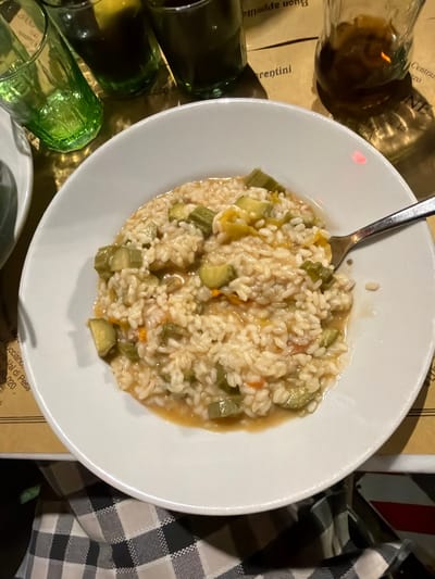 The image captures an overhead shot of a plate of risotto with zucchini, situated on a table setting. The setting suggests a restaurant environment, with glasses of varying sizes, shapes, and hues (brown, green) located to the left of the plate, implying a beverage service. To the right of the plate, a black and white checkered napkin is visible. The table covering has some text in Italian, including the phrase "Buon appetito" which can indicate a city in Italy. The text also includes the city "Val di Pesa," a municipality within the Metropolitan City of Florence in the Italian region of Tuscany. Therefore, the setting can be inferred to be within the vicinity of Florence, Italy. A spoon is resting in the risotto on the plate.