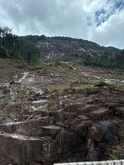 The image depicts a landslide on a steep, rocky mountain slope in Nam Khanh Vinh, Vietnam. The scene is dominated by a vast expanse of exposed rock and soil, with evidence of recent geological activity. The mountain itself is covered in a mix of dense green vegetation and bare rock faces. Streaks of lighter-colored material, likely sediment or mineral deposits, run down the mountain, indicating water flow or past landslides. Scattered rocks and debris are visible across the slope, suggesting a significant upheaval of the terrain. The sky above is overcast with dramatic clouds, adding to the somber atmosphere of the scene. No people or specific structures are identifiable in the image, highlighting the raw power of nature and the impact of the landslide on the natural environment.
