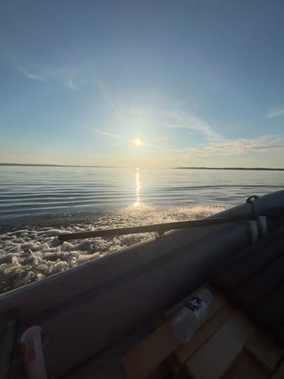 The image shows a serene water scene, likely taken from a boat moving through calm waters. The sun is low in the sky, casting a bright reflection on the water's surface and creating a shimmering path. Wispy clouds are scattered across a clear blue sky. In the foreground, a portion of a boat's interior is visible, including a grey railing and some wooden surfaces. A plastic water bottle with a label is also present inside the boat. The distant horizon shows a faint outline of land. The scene suggests a peaceful moment of travel on a body of water, possibly a lake or a calm sea, under a bright sky. The location is identified as Siukterka, Russia.