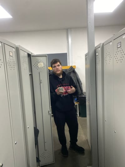 This image depicts an indoor scene, likely a locker room or storage area, within an indeterminate building in the city of Oktyabrskiy, Russia. A single male subject stands facing the camera, positioned between two rows of closed, light-grey metal lockers. He has dark hair, a beard, and is wearing a black hooded sweatshirt with "ORIGINAL BOSTON" printed in red and white on the front, black pants, and black athletic shoes. He holds a small, light-colored rectangular object, possibly a phone or small wallet, in his left hand, while his right hand is raised slightly with fingers curled. The locker immediately to his left is partially open, revealing a dark interior. In the background, to the subject's right, a dark jacket is hanging on a hook against a light-colored wall, and a dark waste bin or container is visible. The floor is dark grey, and the ceiling is white with visible fluorescent light fixtures. A light grey metal structural pole extends vertically from the floor to the ceiling on the right side of the frame.