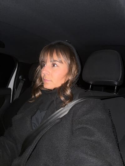 A young woman with bangs and dark curly hair wears a hoodie and sits in a car. She looks down to the left. The interior of the car is dark, with a black headrest and seat to her left. A seatbelt crosses her body. The interior light is off, casting shadows. A glint of exterior light can be seen through a gap in the seat.