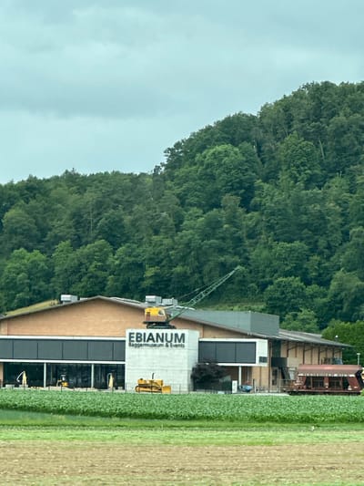 This image depicts the exterior of the EBianum Baggermuseum & Events facility in Zurzach, Switzerland. The building has a modern architectural design with a prominent white section displaying the museum's name. A large green crane is positioned on top of the building, and various construction vehicles, including yellow excavators, are visible in front of the facility. The museum is surrounded by lush green fields and a dense forest covering a hillside in the background. The sky is overcast, suggesting a cloudy day. The scene appears to be a static, daylight capture of the museum and its grounds.