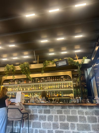 This is a medium shot of an indoor bar area in Dobrota, Montenegro. The perspective is from below eye level, looking up towards the bar and ceiling. The bar itself is made of light-colored brick, with a wooden countertop. Behind the bar, shelves are packed with numerous bottles of liquor, arranged in rows and illuminated from beneath by white LED lights. Green ivy vines hang down from a ledge above the shelves, adding a touch of greenery.

On the left side of the frame, a woman with reddish-blonde hair, wearing a black top and a white skirt or dress, stands at the bar, looking down at a laptop. Her hands are resting on the laptop keyboard. To her right, a light-colored box sits on the bar. To the right of the box, another person with dark hair is visible, appearing to work behind the bar. The background includes more shelves with bottles and some glassware. The ceiling is dark and textured, with several rectangular LED light fixtures casting a bright glow. A black infrared heater is mounted on the ceiling structure above the bar. The overall atmosphere appears to be that of a contemporary bar or restaurant.