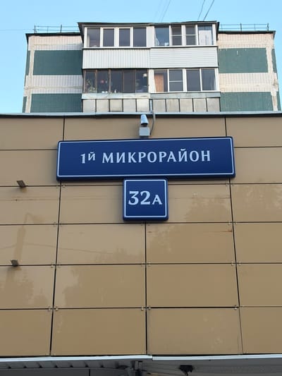 The image shows a building facade in Moskovskiy, Russia, with two blue street signs. The upper sign reads "1Й МИКРОРАЙОН" (1st Microdistrict), and the lower sign reads "32А". The building has a beige tiled facade, and above it, part of an apartment building with green and white horizontal stripes and several windows is visible. The sky is clear and blue. There are no people visible in the image.