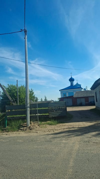 The image shows a street scene with a blue sky and scattered clouds. In the foreground, there is a paved road and a dirt area with a metal fence. Behind the fence, there are trees and buildings, including a blue and white church with a blue dome and cross. There are power lines and a utility pole visible in the upper left portion of the image. The overall scene appears to be in a rural or semi-rural area, possibly in the city of Stowbtsy, Belarus, given the architectural style of the church. No people are visible in the image.