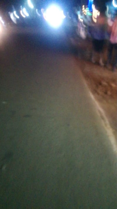 A blurry, low-quality image shows a dark asphalt road extending into the distance. On the right edge, a section of brown dirt or gravel is visible. Further away, blurred figures of people stand on the right side. Several bright, indistinct light sources create large, circular flares in the background, along with streaks of light above the road. The overall scene appears to be captured at night or in very low lighting conditions, resulting in poor focus and significant blurring.