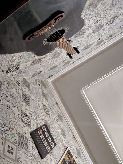 The image shows a dark acoustic guitar laying on a patterned floor. The guitar is angled upwards toward a white wall. The wall has a white trim and is angled at approximately 45 degrees to the floor. In the lower right portion of the image, a small photograph of a man and woman can be seen. The patterned floor is composed of square tiles in various shades of white, blue, black, and gray. The image appears to be from the corner of a room, with the wall and floor meeting at a 90-degree angle. The location is in the city of Lagadar, Indonesia, with coordinates -6.928 latitude and 107.535 longitude.