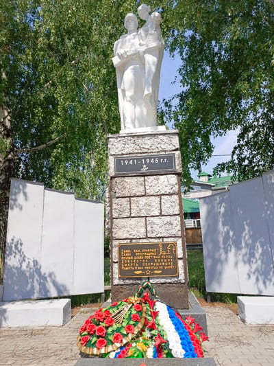 This is a full shot of a war memorial in Zalesovo, Russia, on a bright, sunny day with a clear blue sky and green trees. The memorial features a white stone monument with two figures at the top, possibly representing soldiers or civilians. The base of the monument is made of textured gray stone blocks. On the front of the monument, a dark plaque is inscribed with "1941-1945 rr." and a star symbol. Below this, another gold plaque bears Russian text, which translates to "Glory to you, brave ones! Glory to you, selfless ones! The people sing glory to you, the immortal ones. Death is not the end, you will never be forgotten!" Below the plaque is a large, ornate wreath of red roses and other flowers, with a ribbon of the Russian flag colors (white, blue, and red) at its base. To the left and right of the central monument are two plain, tall, white rectangular structures, also appearing to be part of the memorial. The ground in front of the monument is paved with gray and tan bricks, with some dry twigs scattered on the surface. In the background, a residential building with a green roof is visible, as well as more trees.