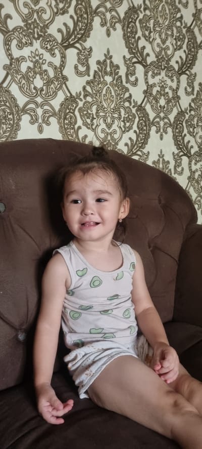 A young girl with dark hair, pulled into a bun, sits on a brown, tufted couch against a backdrop of ornate, damask-patterned wallpaper. She wears a sleeveless white tank top with a pattern of green avocado slices and white shorts. Her head is turned slightly to her right, and she has a happy expression with her mouth open, revealing her teeth. A small pink earring is visible in her right ear. Her left arm rests on her lap, and her right hand is near her leg. The lighting suggests an indoor setting, possibly a home, in Urgench, Uzbekistan.