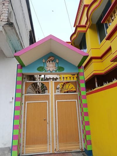 This is a low-angle, full-shot photograph of a brightly painted house entrance in Benapole, Bangladesh. The foreground features a metal gate, divided into two sections, with vertical golden-brown slats. The gate is framed by pillars decorated with a checkerboard pattern of alternating pink and green squares. Above the gate, an archway is painted sky blue and adorned with a relief sculpture of a lotus flower, two lotus leaves, and a conch shell. The archway is capped by a triangular roof structure with pink trim and green undersides.

To the left of the gate, a white wall of a building is visible, showing some signs of wear and tear, including exposed brickwork near the top. To the right, a vibrant yellow building rises up, characterized by curved architectural elements and red trim along its edges and windows. The sky is overcast. The image captures a typical scene of residential architecture in the region.