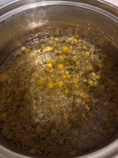 A close-up, top-down view shows the interior of a cylindrical, metallic pot filled with a pale yellow-brown liquid containing numerous chamomile flowers and plant matter. The liquid appears to be hot or simmering, evidenced by condensation droplets visible on the interior rim of the stainless steel pot, particularly prominent on the upper left and right sides. Yellow-orange chamomile flower heads, varying in size, are distributed throughout the liquid, interspersed with lighter, finer plant fragments. The surface of the liquid shows some subtle effervescence or bubbling. No individuals are present. This scene occurs in the city of Voluyak, Bulgaria.