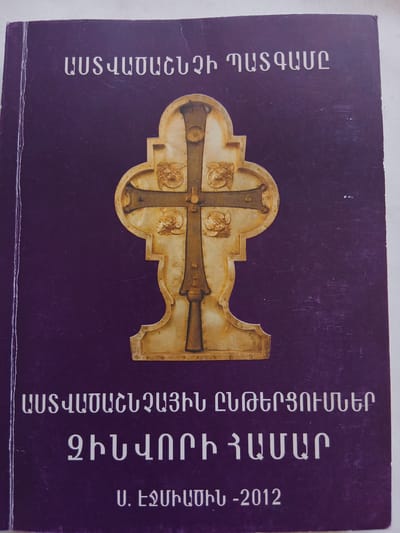The image displays the front cover of a book, predominantly dark purple in color, positioned on a white surface. Centered on the cover is a golden-hued, ornate cross in relief, appearing to be crafted from metal or a similar material. The cross features wide, flaring arms with a central vertical beam, and is adorned with decorative bands and circular elements at its ends and intersection. Each circular element contains a sculpted or embossed figure resembling an angel or cherub. The cross stands on a multi-tiered, sculpted base.

The top of the cover features white Armenian text that reads "ԱՍՏՎԱԾԱՇՆՉԻ ՊԱՏԳԱՄԸ", which translates to "The Message of the Bible" or "The Testament of the Bible".

Below the central cross, additional white Armenian text is present. The first line reads "ԱՍՏՎԱԾԱԲԱՆԱԿԱՆ ԸՆԹԵՐՑՈՒՄՆԵՐ", meaning "Theological Readings". The second line states "ԶԻՆՎՈՐԻ ՀԱՄԱՐ", which translates to "For the Soldier".

At the very bottom of the cover, also in white Armenian script, is "Ս. ԷՋՄԻԱԾԻՆ -2012". This indicates "Holy Echmiadzin - 2012". Holy Echmiadzin is the Mother See of the Armenian Apostolic Church, located in the city of Vagharshapat, Armenia, signifying the publication's origin and year.

The book cover exhibits minor surface abrasions and light scratches, consistent with typical handling and age. No people or other specific objects are depicted on the cover beyond the book itself and the depicted cross.