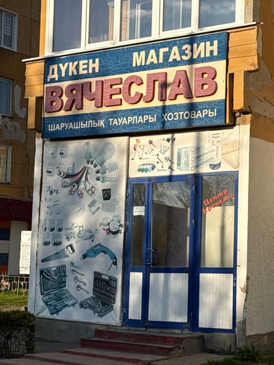 The image depicts the exterior of a hardware store named "Vyacheslav" (Вячеслав) located in Aksay, Kazakhstan. The store's sign features the store's name written in Cyrillic and is accompanied by text in Kazakh and Russian, translating to "Hardware Store".  A poster displays various hardware products like drills, tool sets, light bulbs, and electrical components, covering the storefront entrance. The entrance consists of a blue-framed double door, with partially visible windows and white paneling. A smaller sign with Cyrillic text and a logo is visible in the lower right corner. The building's facade shows signs of weathering, with peeling paint and aging materials. A set of steps lead up to the entrance.