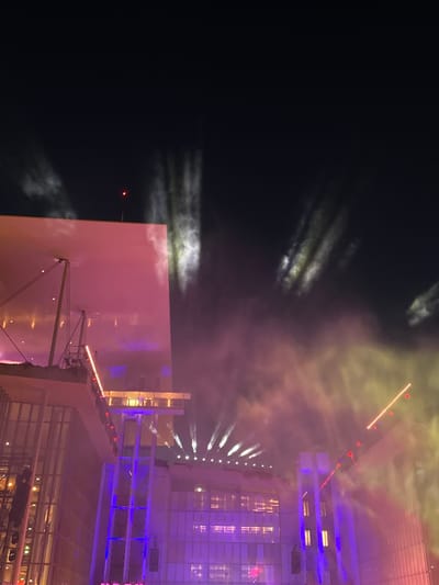 This is a vertical, low-angle, nighttime photograph capturing a dramatic light show or pyrotechnics display above a modern, illuminated building. The scene is dominated by a dark sky filled with wispy clouds and streaks of light. Several beams of light, some appearing white and others yellow or pink, emanate from different points, creating a sense of movement and energy. The building itself is a prominent feature, with multiple levels and a facade that is lit up in shades of purple and yellow. The architecture appears to be contemporary, with large windows and structural elements visible.
The activity taking place is clearly a light show, possibly fireworks or a laser performance, as indicated by the bursts and beams of light in the sky. The building seems to be a venue or a backdrop for this event. The time of day is night, as evidenced by the dark sky and the artificial illumination of the building and lights. There are no visible people or specific location cues beyond the architectural style, but the prompt provides Athens, Greece as context. There is no visible text in the image. The overall mood is one of spectacle and excitement, with the vibrant lights contrasting against the dark sky.
